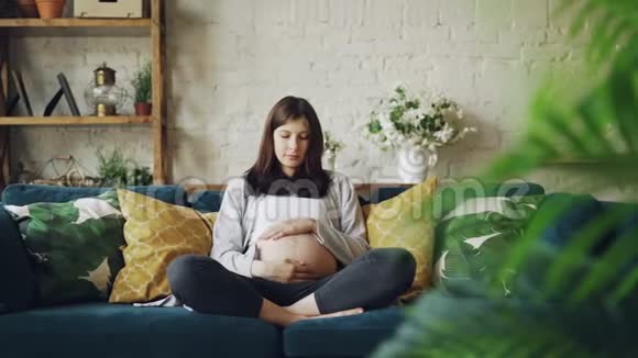 漂亮的黑发女郎期待着母亲抚摸她的大肚子表达对坐在沙发上的胎儿的爱视频的预览图