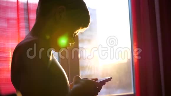 一个年轻人站在窗户附近手里拿着一部日落的时候太阳光线穿过房间视频的预览图