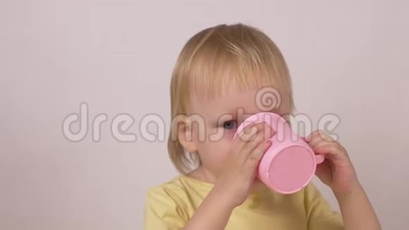 一个金发白种人的小女孩喝着粉红色杯子里的水看着镜头白色背景特写镜头视频的预览图