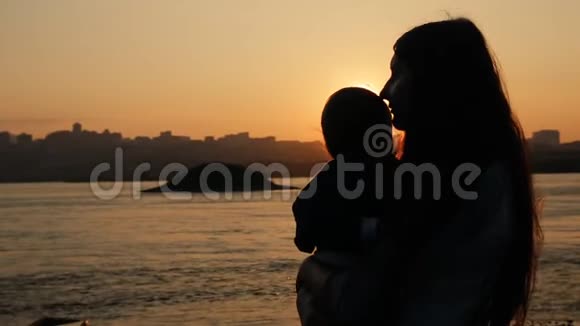 一位年轻母亲在日落时亲吻她站在河边的小儿子的慢镜头视频的预览图