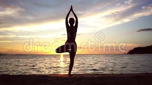 暑假里在日落时分女人在海滩上练习树瑜伽姿势视频的预览图