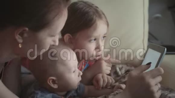年轻的母亲和两个孩子在沙发上玩平板电脑视频的预览图
