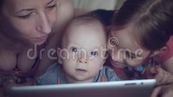 年轻的母亲和两个孩子在沙发上玩平板电脑视频的预览图