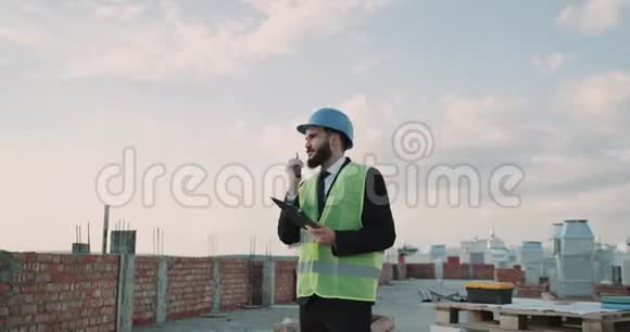 在工地的屋顶上商人戴着安全帽穿着能见度很高的背心用黑色说话视频的预览图