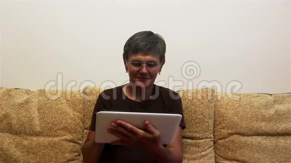 漂亮的成年女性在家里坐在沙发上一边在平板电脑上工作视频的预览图