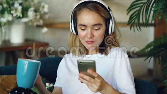 快乐的年轻女士正在使用智能手机在线聊天听音乐无线耳机坐在沙发上视频的预览图
