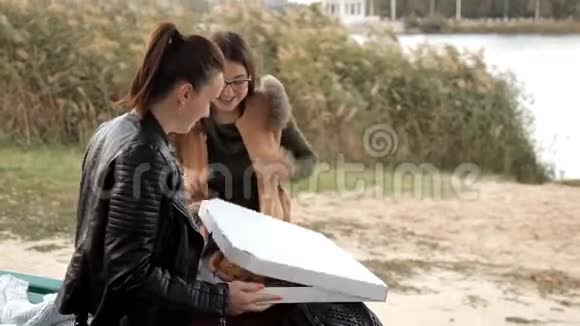 两个女孩坐在河岸上打开一个盒子里面有一个大披萨视频的预览图