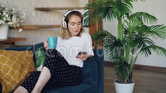 漂亮的女孩正在使用智能手机通过无线耳机听音乐在家坐在沙发上喝茶视频的预览图