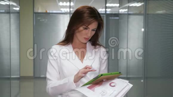 戴着平板电脑和文件的红发女商人站在办公室走廊视频的预览图