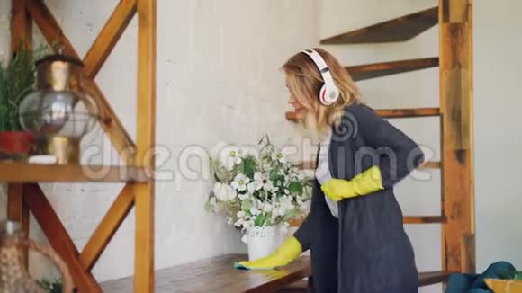 快乐的女人在日常家务中用耳机听音乐跳舞和唱歌女孩在擦桌子视频的预览图