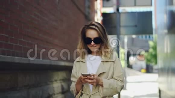 漂亮的金发女孩正在使用现代智能手机触摸屏幕在城市里行走和微笑年轻女子穿着视频的预览图