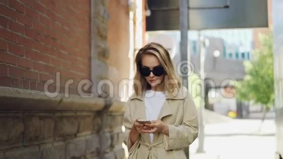 漂亮的年轻女性金发穿着时髦的外套和太阳镜在现代城市里使用智能手机行走视频的预览图
