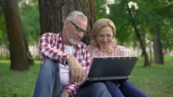 快乐的退休男女坐在草地上在笔记本电脑上看电影视频的预览图