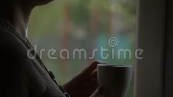 女人喝着一杯热饮料站在窗口透过玻璃看着雨视频的预览图