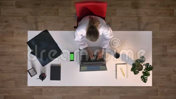 年轻的女科学家穿着白大褂在笔记本电脑上打字打着带有色度的电话头顶镜头坐在X光桌旁视频的预览图