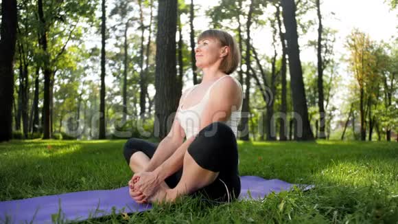 在公园草地上练习瑜伽的40年代女性慢动作视频成年女士在森林里的垫子上做健身运动视频的预览图