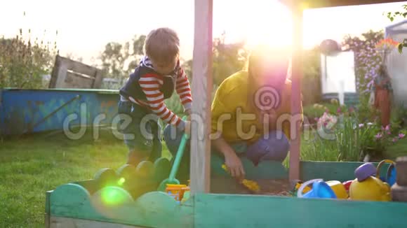 一位年轻的母亲带着一个孩子在沙箱里玩耍夏日操场上的阳光视频的预览图