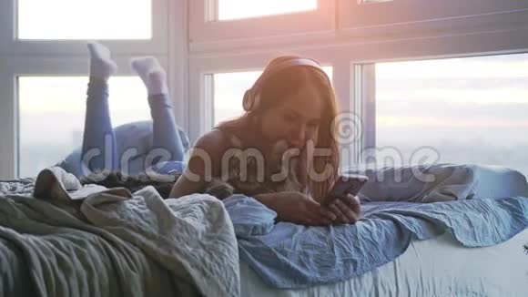 戴着耳机的年轻美女和她可爱的缅因州浣熊猫一起放松听音乐唱歌躺在床上视频的预览图