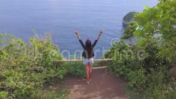 年轻的混合种族旅游女孩站在悬崖边缘与惊人的蓝海伸展的手臂快乐视频的预览图