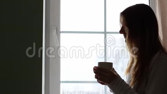 年轻漂亮的女孩正在大窗户附近喝着咖啡透过窗户看着外面视频的预览图