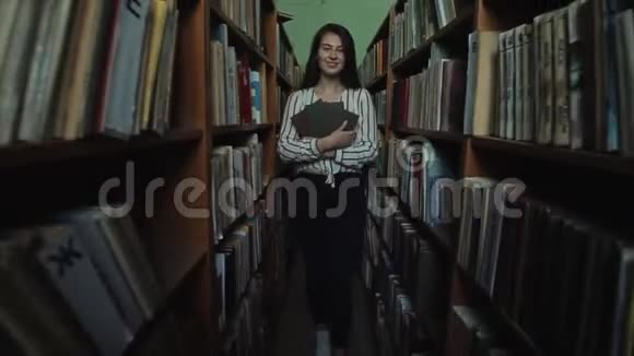 一个留着长长黑发的漂亮女孩走到图书馆的书架之间手里拿着书慢动作广泛视频的预览图