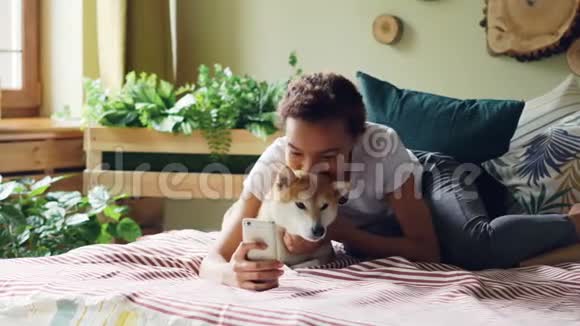 快乐的狗主人正在和可爱的宠物一起躺在床上抱着智能手机摆姿势拥抱动物视频的预览图