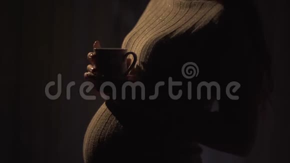 不明身份的怀孕女孩站在一个温暖的毛衣在黑暗的房间里晚上拿着一杯热茶夫人视频的预览图