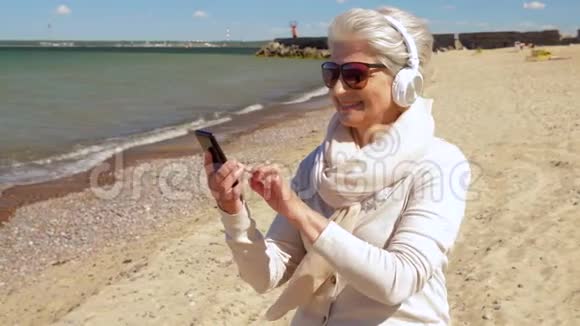 海滩上戴着耳机和智能手机的老太太视频的预览图