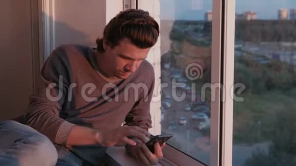 疲惫的男人看着手机在坐在阳台上的社交网络中翻着磁带在日落时分看到城市景色视频的预览图