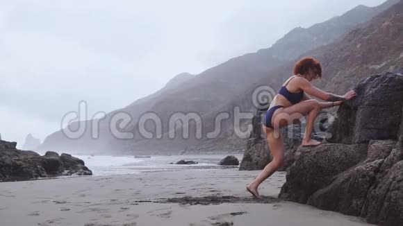 一个女人在靠近海洋的海滩上从一块岩石上进行深蹲和跳跃视频的预览图