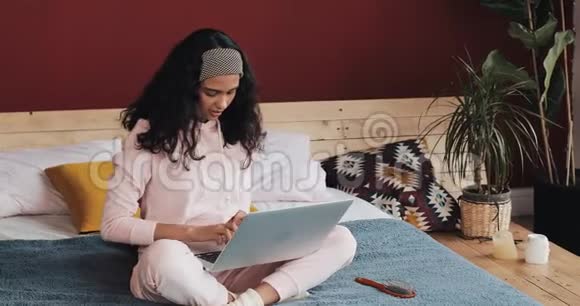 快乐的漂亮女孩穿着粉红色睡衣坐在房子里的床上在笔记本电脑上工作她在微笑视频的预览图