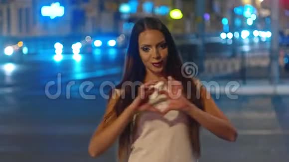 美丽的年轻女子在夜晚的城市展示胜利和心形标志相机上有模糊的交通灯视频的预览图