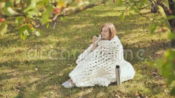一个年轻的女孩裹着一个美利奴格子坐在城市公园的草地上一杯热水瓶里的茶温暖了她视频的预览图