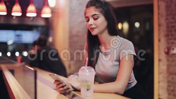 一个女人坐在酒吧里喝着莱曼纳德晚上用她的智能手机十几岁的时髦女孩和她聊天视频的预览图