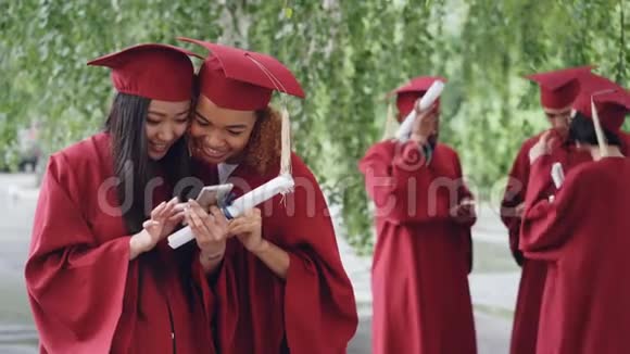 有魅力的年轻女性快乐的毕业生们会在智能手机上观看照片并在毕业当天与她们交谈视频的预览图