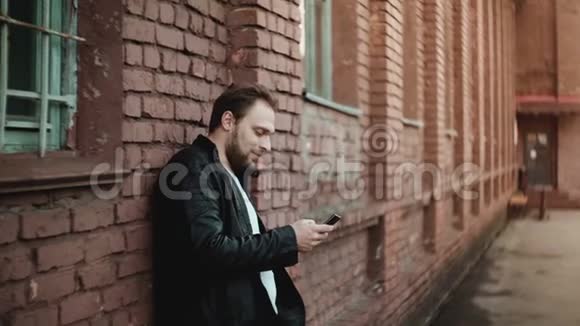 快乐成功轻松的欧洲男人使用智能手机社交应用程序外面和微笑红砖墙的背景视频的预览图