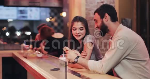 有魅力的年轻夫妇晚上晚些时候在咖啡馆里使用智能手机朋友交流情侣约会聊天视频的预览图