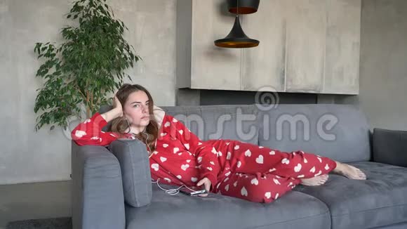 美丽的姜女穿着红色睡衣白天躺在沙发上通过耳机听音乐视频的预览图