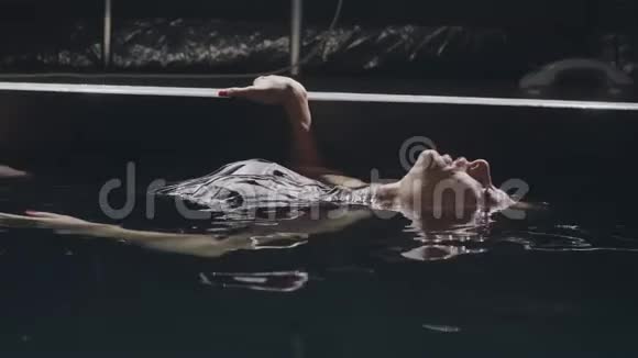 年轻女子躺在黑暗背景下的游泳池里光滑的水面上视频的预览图