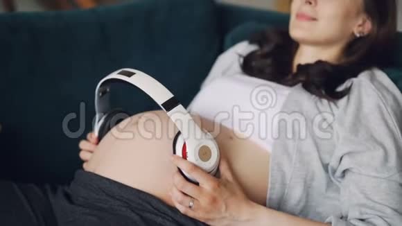 微笑的孕妇躺在舒适的沙发上大肚子上戴着耳机表达对婴儿的爱视频的预览图