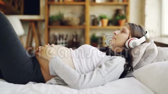 迷人的孕妇正在家里用耳机躺在床上抚摸肚子听音乐闲暇视频的预览图