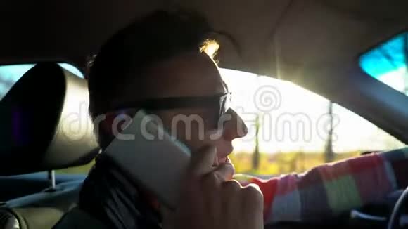 年轻人开着车在户外的日落天空用手机说话特写男伙计戴眼镜的司机视频的预览图