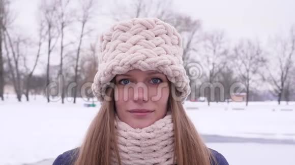 一位戴着白色针织帽子和围巾的蓝眼睛美女站在冬季城市公园里看的画像视频的预览图
