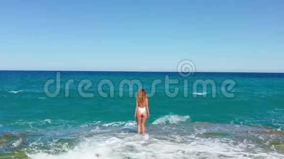 年轻女子穿着比基尼在海边和飞溅的水波空中景观多石海滩无人机上的美女视频的预览图