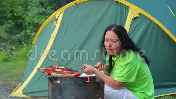 一个穿着绿色T恤的女人坐在森林里的一个帐篷的背景上火在她的胸罩里吹着这就是视频的预览图