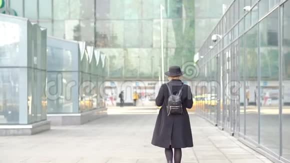 戴着黑色外套帽子的漂亮金发女孩在电话里发短信沿着建筑玻璃窗墙走拍摄慢镜头视频的预览图