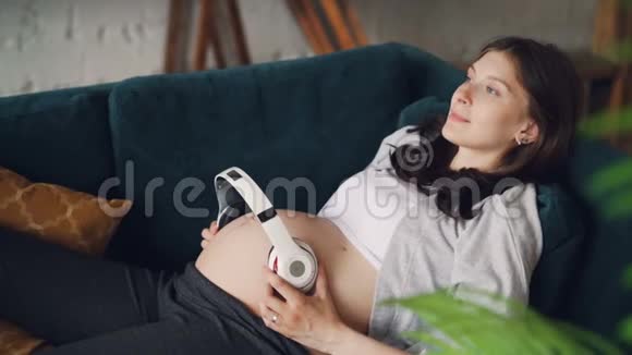 爱期待的母亲抱着无线耳机在她的肚子躺在沙发上在家里休息现代技术视频的预览图