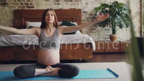 美丽的准妈妈正在现代公寓里练习哈他瑜伽坐在垫子上穿着孕妇运动服视频的预览图