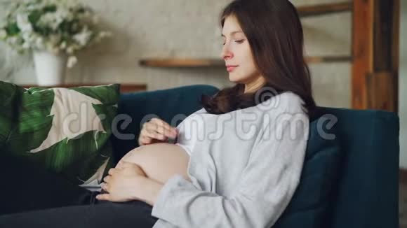 漂亮的怀孕女孩抚摸着她的婴儿肿块带着爱和温柔坐在舒适的沙发上看着它视频的预览图