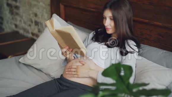 美丽的孕妇正在家里躺在床上看书微笑着享受文学怀孕快乐视频的预览图
